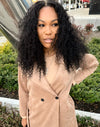 woman wearing e-curly bundles and matching e-curly frontal, wearing a tan coat standing outside in a parking lot in front of bushes