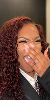 woman with red e-curly bundles wearing a suit jacket, holding her hand up to her face laughing against a black background