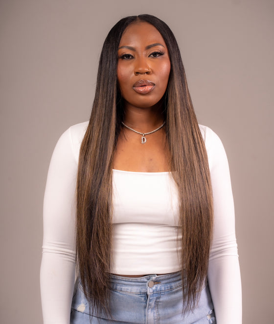 A woman with long straight hair wearing a white top and blue jeans. She has long, straight, balayage colored hair that reaches her shoulders.