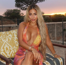  woman seated outdoors wearing multicolored dress and blonde hair extensions, with a backdrop of a sunset.