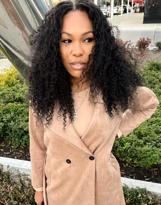 woman wearing e-curly bundles and matching e-curly frontal, wearing a tan coat standing outside in a parking lot in front of bushes