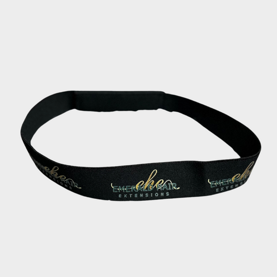 a black elastic hair band with the emerald hair extensions logo shown in a pattern displayed where the band is open in front of a white background.